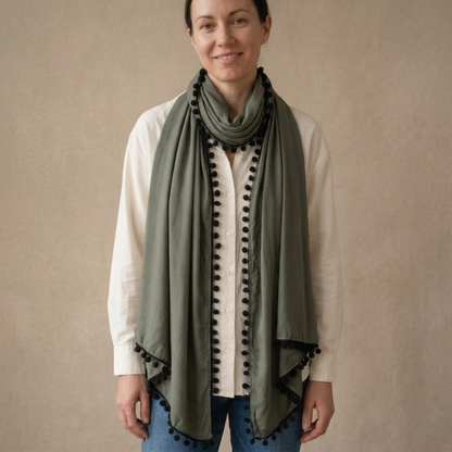 Model wearing a khaki green viscose scarf with black pompom trim, square portrait